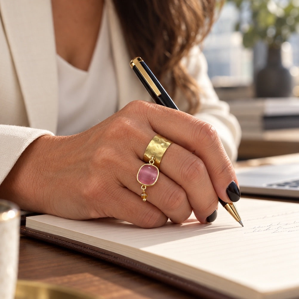 Anello Fru fru a fascia larga in ottone satinato martellato con ciondolo mobile in pietra dura rosa e cristallo color champagne, indossato mentre si scrive su un quaderno