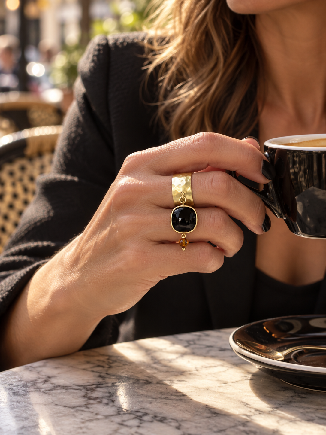 Anello Fru fru a fascia larga in ottone satinato martellato con ciondolo mobile in pietra dura nera e cristallo color champagne, indossato mentre si tiene una tazzina di caffè